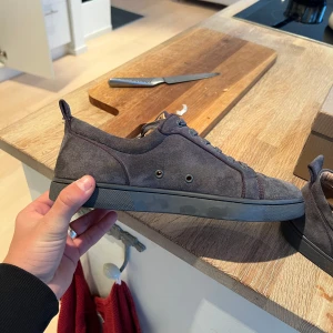 Gråa Louboutin  - Snygga grå sneakers i mocka från Christian Louboutin med den ikoniska röda sulan. Skorna har snörning och en rund tå, vilket ger en stilren look. Perfekta för dig som vill ha en touch av lyx i vardagen.