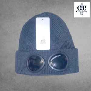CP Company - Snygg mörkblå mössa från C.P. Company med unika glasögondetaljer framtill. Perfekt för att hålla stilen i kallare väder. Ribbstickad design ger en skön passform.