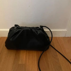 Svart axelväska i syntet - Snygg svart axelväska i syntetmaterial med en minimalistisk design. Väskan har en enkel dragskoöppning och en smidig axelrem. Perfekt för att bära med sig det nödvändigaste med stil.