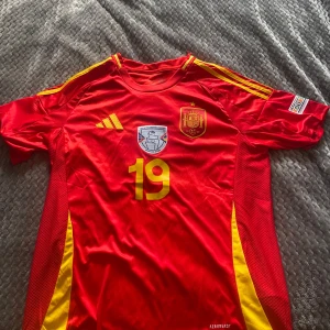 Spanien landslags tröja - Säljer en röd fotbollströja från Adidas med gula detaljer och nummer 19. Tröjan har korta ärmar och Spanien-emblem på bröstet. Perfekt för fotbollsfans!
