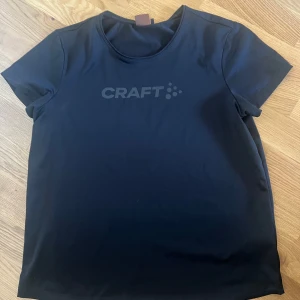 Svart t-shirt från Craft - Säljer en stilren svart t-shirt från Craft. Den har en normal passform och korta ärmar, perfekt för träning eller vardagsbruk. T-shirten är tillverkad i ett syntetmaterial som andas bra. Loggan är diskret placerad på bröstet. 