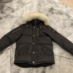 Svart dunjacka med pälsdetalj - Säljer en svart dunjacka från Moose Knuckles med avtagbar pälsdetalj på huvan. Jackan har flera praktiska fickor med dragkedja och knappar, samt en quiltad insida för extra värme. Perfekt för kalla vinterdagar. Inga fel på jackan.