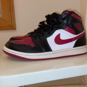Jordan 1 Bred Toe Mid - Snygga Nike Air Jordans i en klassisk färgkombination av svart, röd och vit. Skorna har en hög modell med snörning och den ikoniska swoosh-loggan på sidan. Perfekta för dig som vill ha en stilren och sportig look. (Använda 2-3 gånger)