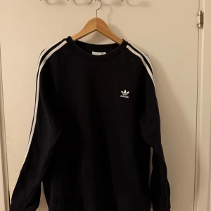 Svart långärmad hoddie från Adidas - Säljer en svart långärmad tröja från Adidas med klassiska vita ränder längs ärmarna och en liten logotyp på bröstet. Perfekt för en sportig look eller avslappnad stil. Tröjan är bekväm och stilren.