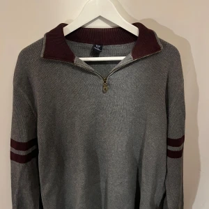 Half Zip hoodie från GAP - Snygg grå tröja från GAP med dragkedja och vinröda detaljer. Tröjan har långa ärmar med två vinröda ränder på ärmarna och en ribbad krage. Perfekt för en stilren och avslappnad look.