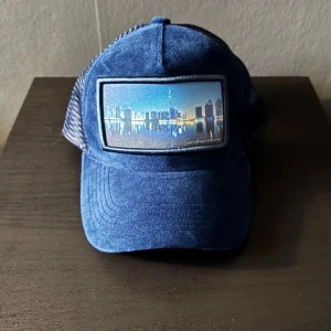 Swedenheadwear keps - Snygg blå keps med ett coolt stadsmotiv på framsidan. Kepsen har ett nätmaterial på baksidan för extra ventilation och en justerbar rem för perfekt passform. Perfekt för en avslappnad stil.