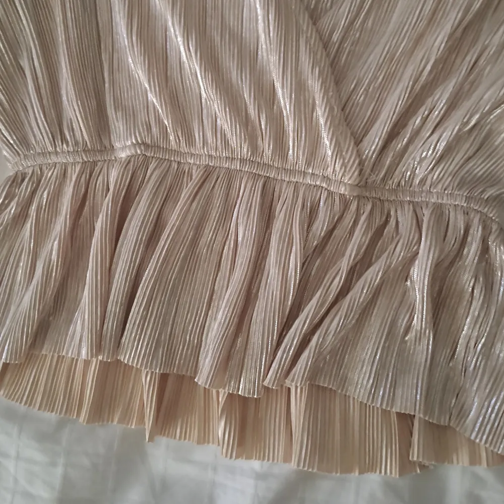Elegant champagnefärgad omlottblus från Gina Tricot. Blusen har långa ärmar och ett glittrigt material som ger en lyxig känsla. Den har en volangdetalj i nederkanten och en v-ringad design.. Puserot.