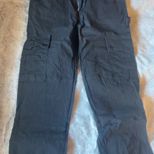 Svarta cargopants med fickor - Säljer ett par svarta cargopants med flera praktiska fickor och knappdetaljer. Byxorna har en loose passform och är perfekta för en avslappnad stil. De är tillverkade i ett slitstarkt material och har en dragkedja framtill.💓
