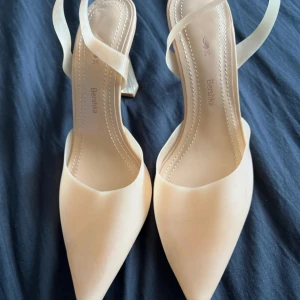 Beige klackskor från Bershka - Snygga beige klackskor från Bershka med spetsig tå och transparent klack. Perfekta för en elegant look. Skorna har en smal rem över foten för extra stil.