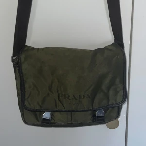 Vintage Prada courier väska - Ovanlig vintage väska perfekt för den som vill sätta en guldkant på sina äventyr på stan. Några små defekter här och där men ingenting med väskans integritet. Alla zip lås fungerar som de ska och väskans interiör är också fullt intakt.