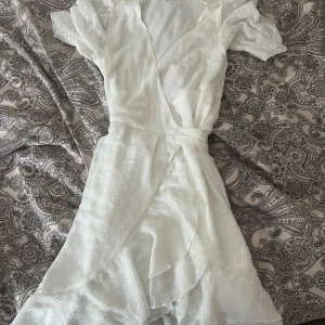 Vit omlottklänning med volangdetaljer - Säljer en söt vit omlottklänning med korta ärmar och volangdetaljer. Klänningen har en knytning i midjan och ett diskret prickigt mönster. Perfekt till konfirmation! Använd endast en gång så den är i nytt skick. Köpt förra året för 699kr.