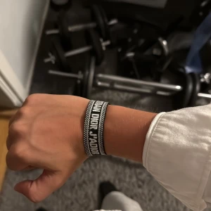 Christian Dior svart/vitt armband - Snyggt armband från Christian Dior i svart och vitt med tydlig logga broderad framtill. Armbandet har justerbar knytning med tofsar i ändarna och är gjort i ett mjukt textilmaterial. Perfekt accessoar för att lyfta din outfit.