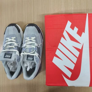 Nike Vomero 5 grå sneakers - Nike Vomero 5 sneakers i olika nyanser av grått och vitt. Ovandel i mesh och syntet med coola detaljer och klassisk Nike-logga på sidan och hälen. Skorna har snörning och en chunky sula som ger en modern vibe. Perfekta för dig som gillar sportig och trendig stil.