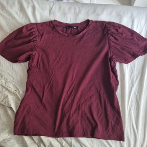 Vinröd topp med puffärm från 157 - Säljer en vinröd topp från 157 med korta puffärmar och rund halsringning. T-shirten är i mjuk bomull och har en enkel, stilren look med en twist tack vare de rynkade axlarna. Perfekt för dig som gillar en basic men ändå trendig vibe.