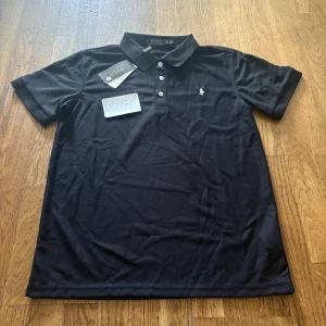 Svart pikétröja från Polo Ralph Lauren - Klassisk svart pikétröja från Polo Ralph Lauren med vit broderad logga på bröstet. Tröjan har korta ärmar, krage och tre knappar framtill. Perfekt för en clean och stilren look. Tillverkad i mjuk bomull som känns skön mot huden.