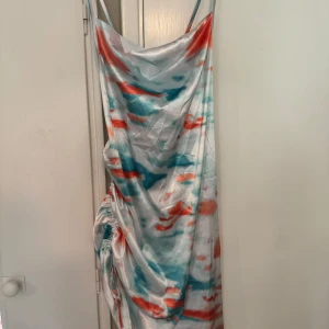Färgglad satin klänning med snörning - Säljer en unik klänning i satin med färgstarkt mönster i turkos, orange och vitt. Klänningen har smala axelband, draperad framsida och snygg snörning längs ena sidan för en cool effekt. Går till låren. Perfekt för dig som vill sticka ut med en statement piece!