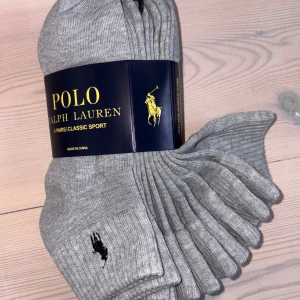 Gråa ribbade strumpor Polo Ralph Lauren - Sexpack klassiska gråa ribbade strumpor från Polo Ralph Lauren med svart broderad logga vid ankeln. Perfekta till sneakers eller loafers och tillverkade i mjuk bomullsblandning för skön passform hela dagen.