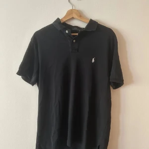 Svart pikétröja från Ralph Lauren - Klassisk svart pikétröja från Ralph Lauren med vit broderad logga på bröstet. Tröjan har korta ärmar, krage och två knappar framtill. Tillverkad i mjuk bomull som känns skön mot huden. Perfekt för en clean och stilren look. Matrailet  är skönt inte som det flesta piké tröjorna  perfekt till sommaren 