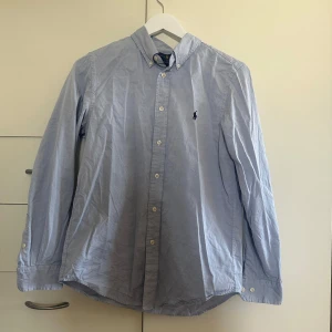 Ljusblå skjorta från Ralph Lauren - Klassisk ljusblå skjorta från Ralph Lauren med button-down krage och broderad logga på bröstet. Skjortan har lång ärm, vita knappar och är i bomull. Custom fit-modell som ger en snygg passform. Perfekt till jeans eller chinos.