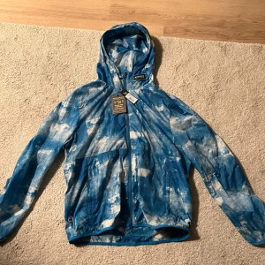 Blå mönstrad vindjacka från Woolrich - Säljer en blå och vit vindjacka från Woolrich med coolt abstrakt mönster. Jackan har huva, hel dragkedja och två fickor framtill. Perfekt för dig som vill sticka ut med en unik look. Lätt och smidig att bära, passar till många outfits.