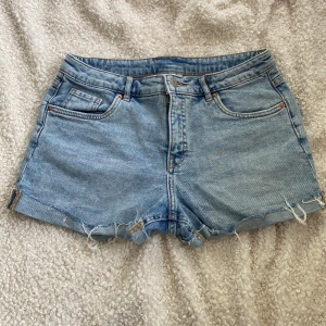 Ljusblå jeansshorts från Kappahl - Snygga ljusblå jeansshorts från Kappahl med råa, fransiga kanter och klassisk femficksdesign. Shortsen har normal passform och dragkedja framtill. Perfekta för sommardagar och chill häng utomhus.