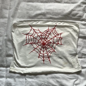Vit tubtopp med rött spindelnät SHEIN - Cool vit tubtopp från SHEIN i stretchigt material med ett stort rött spindelnätsmotiv framtill. Toppen är bandeau-modell och har en enkel, tight passform. Perfekt för dig som vill sticka ut med en edgy detalj i din outfit. Tyget är något genomskinligt och tröjan har en sminkfläck på sidan💞