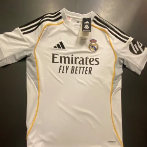 Real Madrid matchtröja Adidas 13-14 år - Snygg vit Real Madrid fotbollströja från Adidas med svarta axelränder, gula detaljer och klubbmärke på bröstet. Tryck med 'Emirates Fly Better' på framsidan. Tillverkad i lätt funktionsmaterial för bästa komfort på planen. Tar tid för leverans