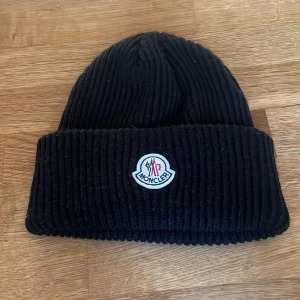 Svart ribbstickad mössa från Moncler - Svart ribbstickad mössa från Moncler med klassisk uppvikt kant och broderad logotyp framtill. Snygg och enkel design som passar perfekt till kalla dagar. Tillverkad i mjukt material som håller värmen och ger en clean look.