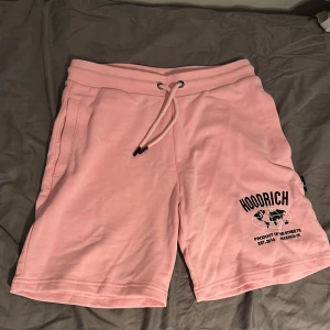 Rosa Hoodrich shorts med tryck - Snygga rosa shorts från Hoodrich med svart tryck och världskarta på ena benet. De har elastisk midja med snörning och sidofickor. Perfekta för chill dagar eller streetwear-stil. Mjukt material som känns skönt mot huden.