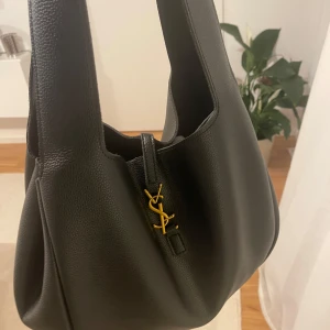 Svart axelväska  - Snygg svart axelväska i lyxigt skinn med guldigt YSL-emblem framtill. Väskan har bred axelrem, rymligt huvudfack och innerficka med dragkedja. Perfekt för dig som vill ha en stilren och lyxig väska med fin  design.