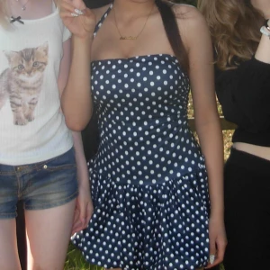 Marinblå prickig kort klänning - Söt marinblå kortklänning med vita prickar och halterneck. Klänningen har en figurnära överdel och utställd kjol som ger en lekfull retrovibe. Perfekt för dig som gillar klassiska mönster och vill sticka ut lite extra.