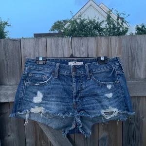 Blå jeansshorts från Hollister - Super snygga blå jeansshorts från Hollister med slitna detaljer och fransig kant som inte passar mig 😭. Klassisk femficksmodell med låg midja och råa kanter för en avslappnad vibe. Perfekta för sommardagar och festivaler. Ikonisk Hollister-logga på knappen och bakfickor med dekorativa sömmar. Storlek W27 men jag skulle säga storlek M då det inte passar mig som bär S