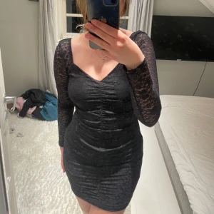 Svart spetsklänning med lång ärm - Säljer en svart kort klänning med figurnära passform och långa ärmar i spets. Klänningen har v-ringning och rynkade detaljer framtill som ger en snygg siluett. Bara använd en gång så är som ny!😍