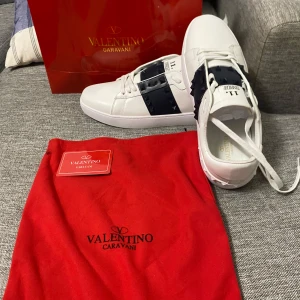 Valentino Garavani Rockstud sneakers - Snygga vita Valentino Garavani Rockstud sneakers med svart band och ikoniska nitar. Skorna har rund tå, snörning och är tillverkade i lyxigt skinn. Perfekt för dig som vill sticka ut med en exklusiv och modern look.