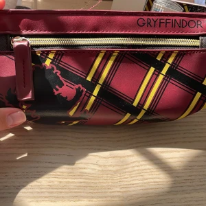 Gryffindor rutig necessär Harry Potter - Necessär med Gryffindor-tema från Harry Potter. Djup vinröd färg med rutigt mönster i gult och svart samt Gryffindor-lejonet tryckt på sidan. Tillverkad i syntetmaterial med dragkedja upptill och praktiskt handtag. Perfekt för att förvara smink eller småprylar.