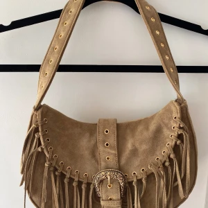 Beige mocka axelväska med fransar - Säljer en beige axelväska i mocka med snygga fransar och guldfärgade detaljer. Väskan har en bred axelrem med nitar och en dekorativt mönstrad spännesdetalj framtill. Perfekt för dig som gillar boho-stil. Endast använd en gång en kort stund, som ny! köpt för 289kr