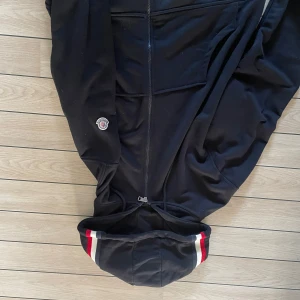 Svart Moncler vindjacka med huva - Svart vindjacka från Moncler med hel dragkedja, huva och snygga röda och vita ränder på huva och ärmslut. Jackan har Moncler-logga på ena ärmen och praktiska fickor framtill. Perfekt för dig som gillar stilren och sportig look.
