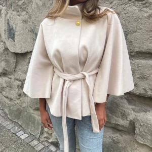 Missäy poncho  - Stilren beige kappa med knytbälte i midjan och vida trekvartsärmar. Hög krage med en dekorativ guldig knapp upptill. Perfekt för dig som gillar en clean och elegant look.