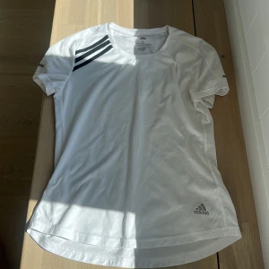 Vit Adidas t-shirt AEROREADY S - Vit t-shirt från Adidas med klassiska svarta ränder på axeln och diskret logga nertill. Modellen är kortärmad och tillverkad i lätt, ventilerande polyester med AEROREADY-teknologi, perfekt för löpning eller träning. Snygg och sportig passform.