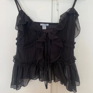 Svart volangig linnetopp från newyorker - Supersöt svart linnetopp från newyorker med tunna axelband, volanger och plisserat tyg. Toppen har knytning framtill och är gjord i ett lätt, transparent material som ger en luftig känsla. 