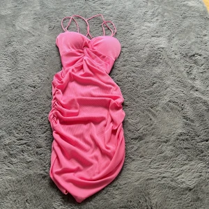 Rosa ribbad kortklänning med smala band - Supersöt rosa kortklänning från 8F Moda med smala axelband och ribbad struktur. Klänningen har rynkade sidor och figurnära passform, perfekt för dig som vill sticka ut. Tillverkad i stretchig polyesterblandning för en bekväm känsla. Klänningen har insydd vadering vid bysten och en korsad rygg.