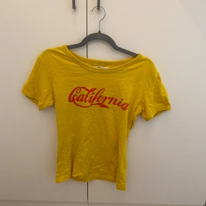 Gul t-shirt med röd California-tryck - Gul t-shirt med coolt rött California-tryck framtill. Modellen är kortärmad och har en avslappnad passform med rund halsringning. Perfekt för dig som vill ha en färgklick i garderoben och gillar retrovibbar.