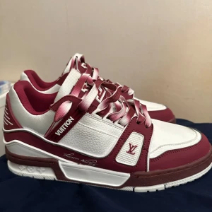 Louis Vuitton sneakers röd/vit 8.5 - Snygga Louis Vuitton sneakers i vitt och vinrött med klassiska LV-detaljer och logga på sidan. Skorna har en chunky sula, rund tå och coola tvåfärgade snören. Tillverkade i skinn och textil för en lyxig känsla. Perfekta för dig som vill sticka ut med exklusiv streetstyle.