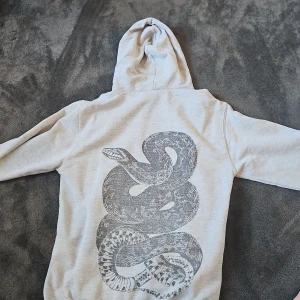 Ljusgrå hoodie The Cool Elephant ormtryck - Säljer en ljusgrå hoodie från The Cool Elephant med stor svart ormtryck på ryggen och liten logga framtill. Tröjan har huva och känguruficka, perfekt för dig som gillar unika prints och streetstyle.