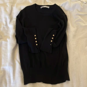 Svart tröja med guldknappar från Zara - Svart långärmad tröja från Zara med snygga guldknappar på ärmsluten. Tröjan har en klassisk rund halsringning och är i mjukt material som sitter skönt på kroppen. Perfekt för dig som gillar stilrena detaljer och enkel design.