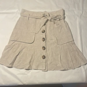 Beige linnekjol med knappar från ASOS - Supersöt beige kjol från ASOS. I linne med stora knappar framtill, fickor och ett brett knytskärp i midjan. Kjolen har volangdetaljer nertill på framsidan. En underbar kjol till sommar garderoben
