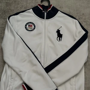 Vit Ralph Lauren hoodie  - Vit hoodie från Ralph Lauren med USA Olympic Team-märke på bröstet och.           stor U.S.A.-text på ryggen. Hoodie har marinblå detaljer längs ärmarna,               ribbade muddar med röd och vit rand,  dragkedja framtill och klassisk sportig            look. Material: 85% bomull, 15% arkul.