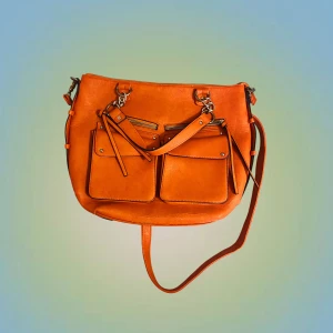 Orange axelväska med dragkedjor - Snygg orange axelväska i skinnimitation med två stora fickor framtill, flera dragkedjor och justerbar axelrem. Väskan har ljus insida med praktiska innerfack och detaljer i silverfärgad metall. Perfekt för dig som vill ha plats för allt du behöver.