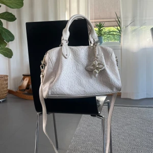 Vit handväska med mönster och berlocker - Snygg vit handväska med präglat blommönster och två blom-charms från märket Liu-Jo!! Väskan har både handtag och axelrem, samt dragkedja upptill. Insidan har flera fack. Denna väskan är liten och smidig (ca 30x15x20) men tro mig när man får plats med ALLT!! 