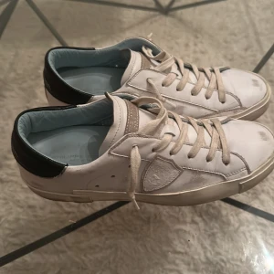 Vita Philippe Model sneakers i skinn - Snygga vita sneakers från Philippe Model med svarta detaljer på hälen och ljusblå insida. Skorna har klassisk snörning, rund tå och är tillverkade i skinn. Perfekta för dig som gillar stilrena och trendiga sneakers med exklusiv känsla.använt dem 3 gånger nypris 3500kr